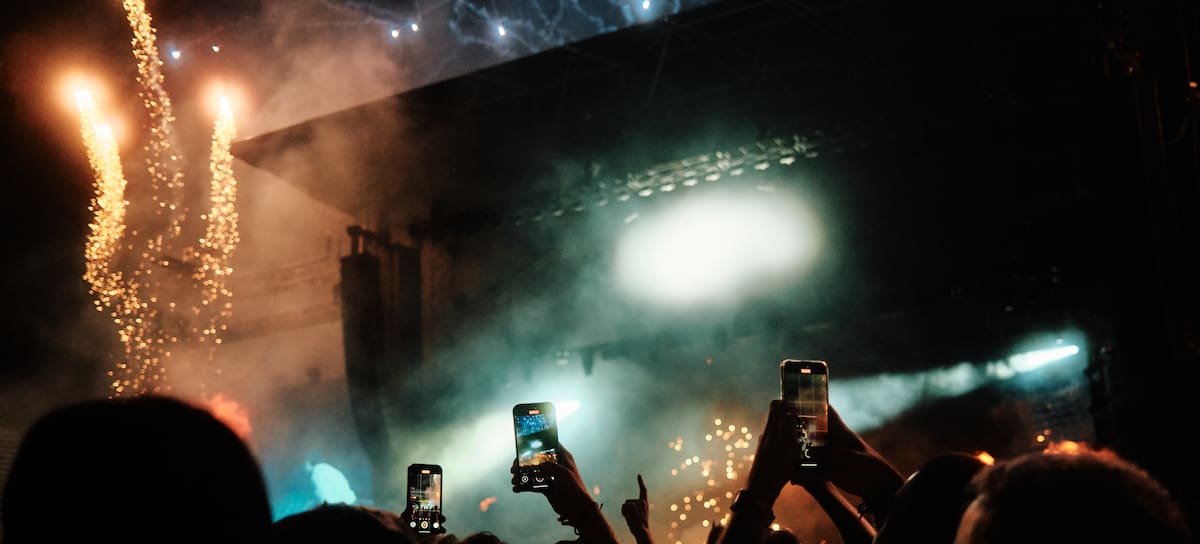 Concert crowd recording the show with smartphones as fireworks and stage lights illuminate the night sky during a live music performance.