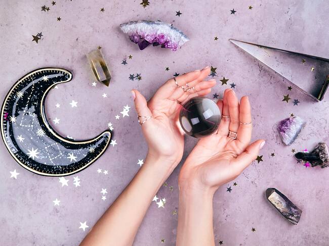 Magic crystals for rituals. Hands with rings on fingers are holding crystal ball near esoteric set on concrete gray background with many stars sequins. Flat lay style