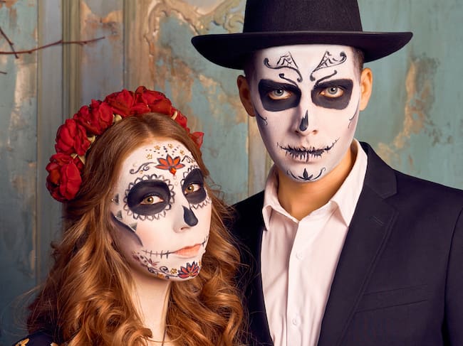 Indoor shot of serious romantic couple pose before Halloween event. People preparing for the popular carnival, dressed in costume, applied makeup in the shape of skull. Day of dead holiday in Mexico.